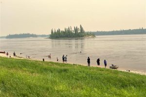 A group of people stand and swim along a grassy riverbank, with a small forested island visible across the calm water under a hazy sky.